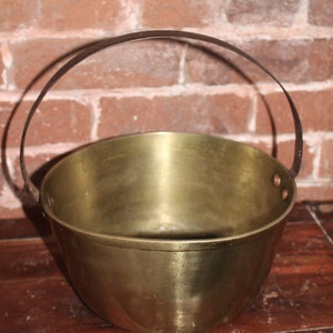 May include: A brass bucket with a black metal handle. The bucket is round and has a smooth, shiny surface.