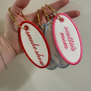 May include: Embroidered oval keychains with gold-tone hardware. One keychain has a red border and the text "smokeshow" in red script. Another has a pink border with "scottie's mom" in pink script. A third keychain has a blue border with text.