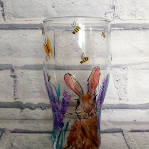 Puede incluir: Un vaso de pinta de vidrio transparente con un dise&ntilde;o pintado a mano de un conejo marr&oacute;n rodeado de flores moradas y mariposas amarillas. El vaso tiene un fondo redondeado y una parte superior recta.