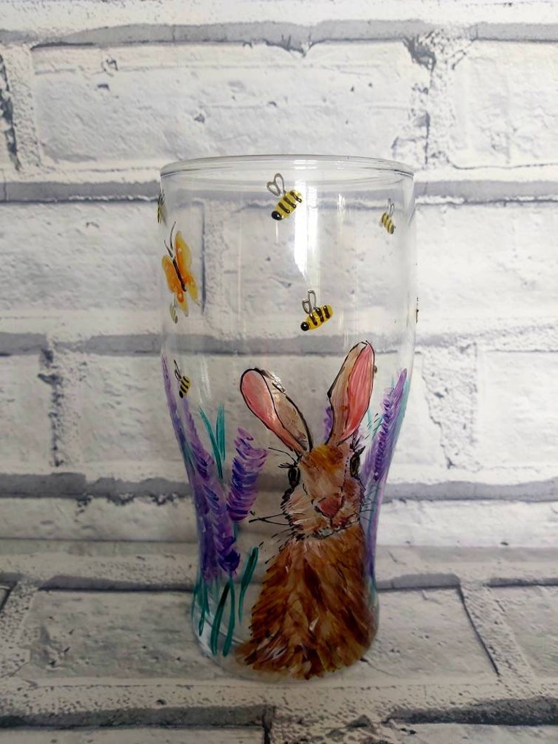 Puede incluir: Un vaso de pinta de vidrio transparente con un dise&ntilde;o pintado a mano de un conejo marr&oacute;n rodeado de flores moradas y mariposas amarillas. El vaso tiene un fondo redondeado y una parte superior recta.