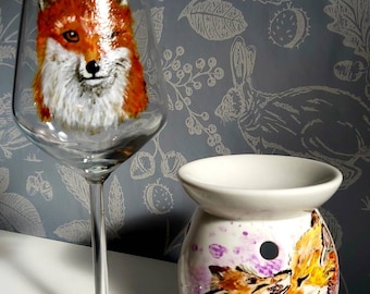 Puede incluir: Una copa de vino de cristal transparente con una cara de zorro pintada a mano en naranja y blanco, y un calentador de cera de cerámica blanca decorado con una escena pintada de dos zorros y acentos de acuarela morada en el cuerpo.