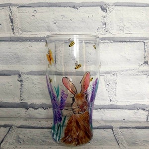 Puede incluir: Un vaso pintado a mano con un conejo marr&oacute;n rodeado de flores moradas y abejas amarillas. El vaso tiene un fondo transparente y es perfecto para una bebida.
