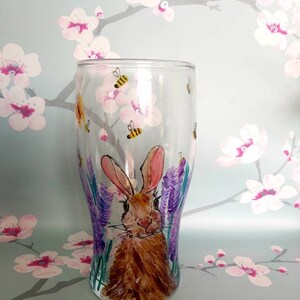 Puede incluir: Un vaso pintado a mano con un conejo marr&oacute;n en un campo de flores moradas. Hay abejas amarillas y negras volando alrededor del conejo.