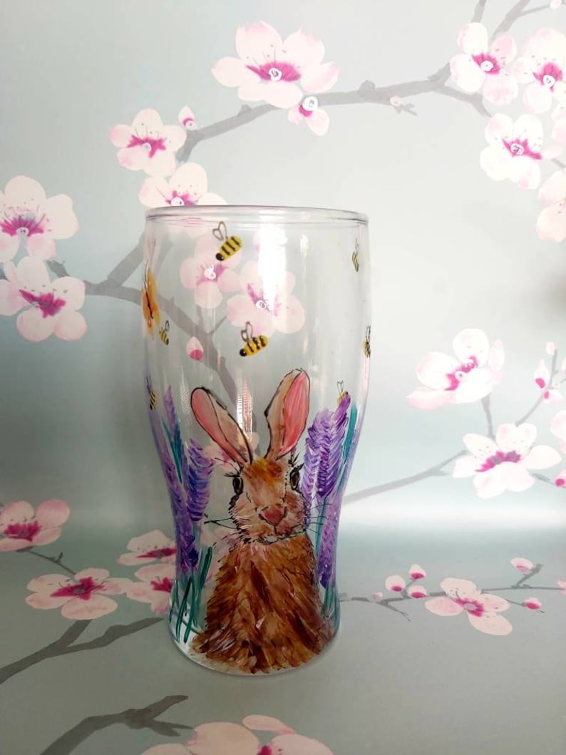 Puede incluir: Un vaso pintado a mano con un conejo marr&oacute;n en un campo de flores moradas. Hay abejas amarillas y negras volando alrededor del conejo.