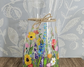 Puede incluir: Un jarrón de vidrio transparente con un lazo de cuerda rústica alrededor del cuello. El jarrón está pintado a mano con un diseño de flores silvestres de colores, que incluye flores amarillas, rojas, rosas, azules y blancas.