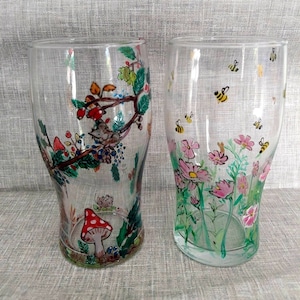 May include: Two clear glass tumblers with hand-painted designs. One tumbler features a woodland scene with a mushroom, a mouse, and a tree branch with berries. The other tumbler features a floral design with pink flowers and green leaves.