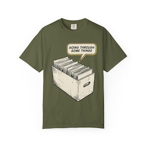May include: Olive green t-shirt featuring a comic-style illustration of a box filled with files. A speech bubble above the box reads "GOING THROUGH SOME THINGS". The design is in black and white with a yellow outline.