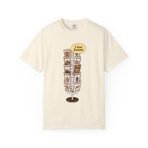 May include: Cream-colored t-shirt featuring a brown and yellow graphic of a postcard display with the text "I Got Issues" in a yellow speech bubble. The shirt has a crew neck and short sleeves.