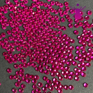 May include: A scattering of small, round, fuchsia-colored rhinestones. The gemstones are scattered across a dark surface, with a few individual stones visible. The rhinestones have a faceted cut, reflecting light and adding sparkle.
