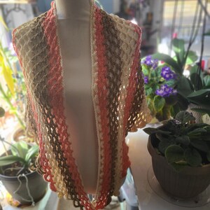 May include: A handmade crochet shawl in shades of peach, cream, and brown, displayed on a mannequin. The open weave design and scalloped edges add a delicate touch to this wearable accessory. This shawl is perfect for adding a layer of warmth and style.