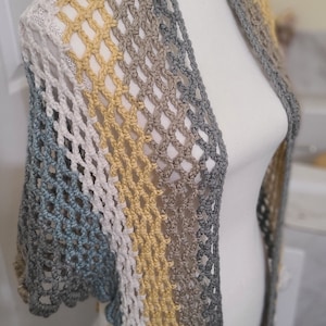 May include: A crocheted shawl with a multi-colored striped pattern in shades of gray, white, and yellow. The shawl is draped over a white mannequin.