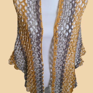 May include: A handmade, open-weave crochet shawl in shades of gray, white, and mustard yellow. The shawl is draped over a white mannequin, showcasing its open front design and scalloped edges. This garment is a stylish accessory.