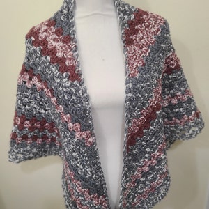 May include: A crocheted shawl in shades of gray, white, and pink. The shawl is made with a textured stitch and has a triangular shape.