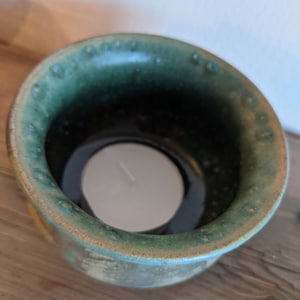 Könnte beinhalten: Ein grüner Kerzenhalter aus Keramik mit einem weißen Teelicht darin. Der Halter hat eine gesprenkelte Glasur und eine abgerundete Form.