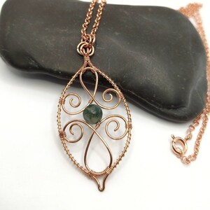 May include: A copper-coloured wire-wrapped pendant necklace featuring a green stone bead. The pendant has an open, leaf-like design with swirling details. The necklace chain is also copper-coloured and has a lobster clasp.