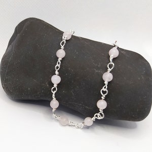 May include: A delicate bracelet featuring pale pink, round beads strung together with silver-coloured wire. The wire is intricately looped between the beads, creating a decorative chain. The bracelet is displayed on a dark, textured stone.