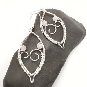 May include: A pair of silver wire earrings in a leaf design. Each earring has a teardrop shape with a woven wire pattern, two small pink beads, and a decorative swirl. The earrings are displayed on a black stone.