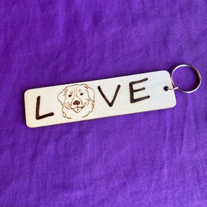 May include: Wooden keychain with a dog's face and the word "LOVE" engraved on it. The keychain has a silver ring.