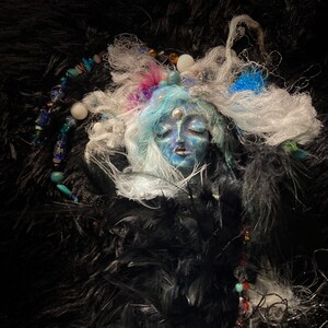 May include: A blue and white face sculpture with closed eyes, surrounded by white and black feathers and beads. The sculpture is set against a black background.