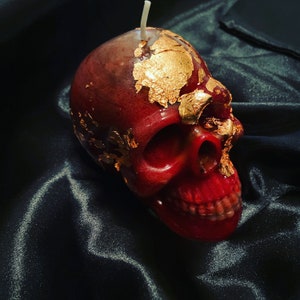 May include: A red skull-shaped candle with gold leaf accents. The candle is sitting on a black fabric surface.
