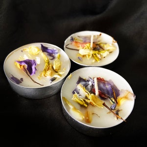 May include: Three white tea light candles with dried flowers and herbs embedded in the wax. The flowers are purple, yellow, and white.