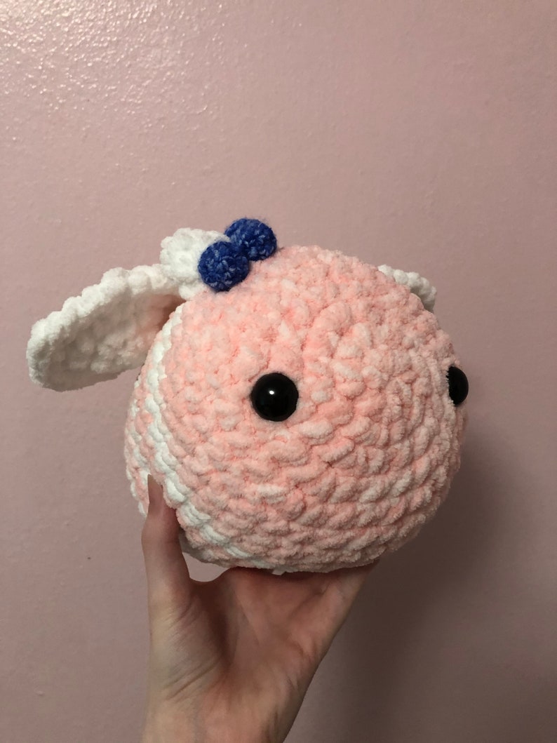 Pu&ograve; includere: Un giocattolo di peluche rosa e bianco all'uncinetto con pompon blu in cima e occhi neri a bottone.