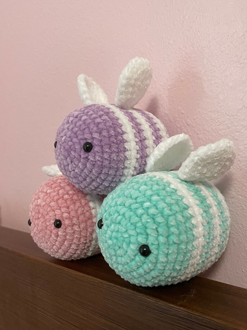 Pu&ograve; includere: Tre peluche di api all'uncinetto in rosa, viola e turchese. Ogni ape ha strisce bianche e ali bianche.