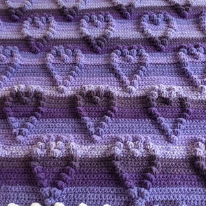 May include: A purple and white crocheted blanket with a heart pattern. The blanket is made with a variety of shades of purple yarn.