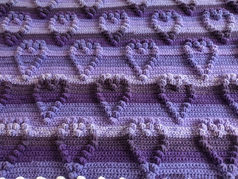 May include: A purple and white crocheted blanket with a heart pattern. The blanket is made with a variety of shades of purple yarn.