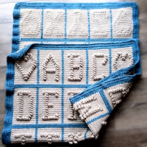 Könnte beinhalten: Eine gehäkelte Babydecke in Blau und Beige. Die Decke zeigt das Wort "MASTER" in erhabenen Buchstaben.