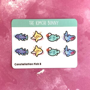 May include: A sheet of stickers featuring a constellation fish design. The stickers are blue, yellow, and green. The text "The Kimchi Bunny" and "Constellation Fish B" are printed on the sheet.