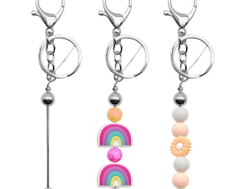 May include: Three silver keychains with different charms. The first keychain has a silver bead and a long silver rod. The second keychain has a silver bead, a glittery orange bead, a pink and blue rainbow charm, a pink glitter bead, and another pink and blue rainbow charm. The third keychain has a silver bead, a white bead, a peach bead, a white bead with a peach flower, a peach bead, and a white bead.