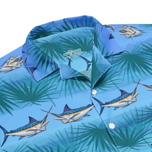 Puede incluir: Una camisa de manga corta con estampado azul y verde con cierre de botones blancos. La camisa presenta un patrón repetido de peces marlin y hojas de palma. La camisa tiene el texto "COYOTES" en blanco sobre fondo negro.