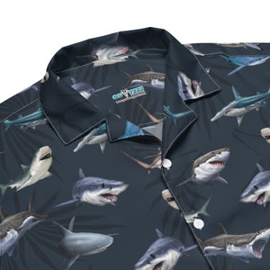May include: A dark blue button-up shirt with a repeating pattern of various shark species. The shirt has a white collar and a single button closure. The text "GO YEEES" is printed on the shirt.