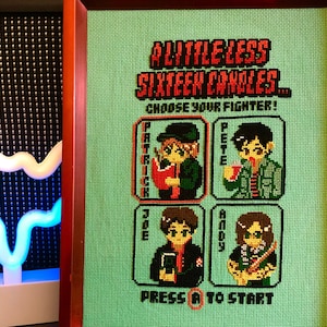 May include: A framed cross-stitch artwork featuring a pixelated video game-style design. The title reads "A Little Less Sixteen Candles... Choose Your Fighter!" with four character options: Patrick, Pete, Joe, and Andy. The bottom of the artwork reads "Press A to Start."