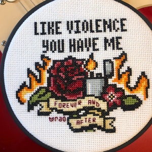 Könnte beinhalten: Ein weißer Stickrahmen mit schwarzem Rand, der ein Kreuzstich-Design mit einer roten Rose, einem Feuerzeug und Flammen zeigt. Der Text "Like violence you have me" befindet sich über dem Design, und "Forever and ever after" darunter.
