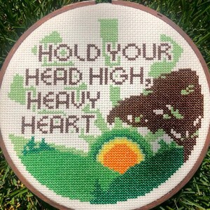 May include: A round embroidery hoop with a white fabric background. The fabric is stitched with green, brown, and orange thread to create a landscape scene with a sun and the words "Hold Your Head High, Heavy Heart".