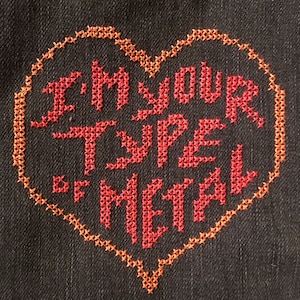 Może przedstawiać: Wzór haftu krzyżykowego w kształcie serca na ciemnym denimie. Wzór jest wykonany czerwoną i pomarańczową nicią i brzmi "I'm your type of metal".