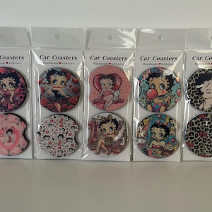 May include: Set of car coasters featuring various Betty Boop designs. Each coaster has a different image of the cartoon character, with text that reads "Car Coasters Handmade with love". The coasters are packaged in clear plastic.