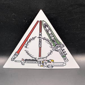 May include: White triangular ceramic tile featuring a design with red and green lightsabers, a brown staff, and a blaster. The design includes a ring of text in a circular pattern. The tile is a decorative item.