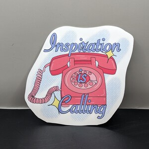 May include: A white sticker with a pink rotary phone and the text "Inspiration is Calling" in blue lettering. The phone has a white dial and a white cord.
