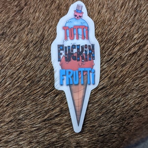 May include: A sticker of an ice cream cone with the words "Tutti Fuckin Frutti" in red, white, and blue. The ice cream cone is topped with a small American flag.