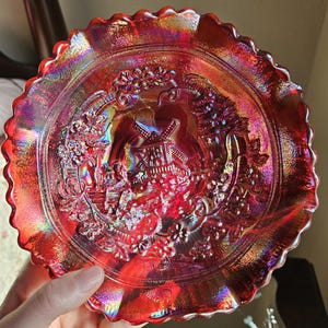 May include: A red iridescent glass plate with a raised design of a windmill and grape vines. The plate has a scalloped edge.