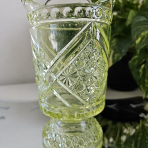 May include: A pale green, vintage-style glass candle holder with a decorative, geometric pattern. The candle holder has a flared top with a handle and a round base. The glass has a diamond and starburst design. The background includes green foliage.