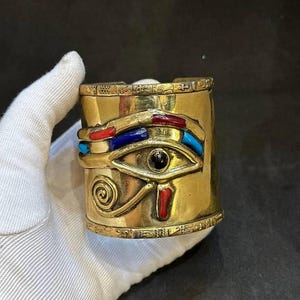 Puede incluir: Un brazalete dorado con un diseño de ojo de Horus egipcio. El ojo está incrustado con piedras azules, rojas y negras.