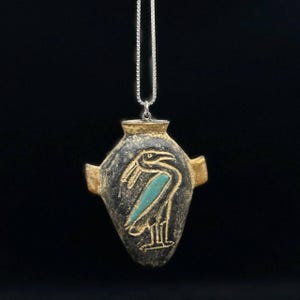 Puede incluir: Un collar con colgante con un diseño del antiguo Egipto. El colgante tiene forma de jarrón gris oscuro con borde y detalles dorados. Una figura de pájaro turquesa está representada en la parte delantera. La cadena del collar es plateada.