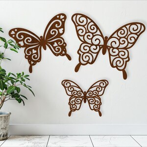 May include: Three brown metal butterfly wall decor pieces with intricate designs. The butterflies are different sizes and are arranged in a staggered pattern on a white wall.