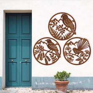 May include: Three circular, brown metal wall art pieces featuring bird and mountain designs. Each piece has a different bird perched on a branch with foliage, set against a mountain backdrop. The art is mounted on a white wall.
