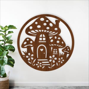 May include: A brown wooden mushroom house wall art piece. The design features a house with a heart-shaped door, windows, and a chimney, surrounded by smaller mushrooms and flowers. The art is cut out in a circular shape.