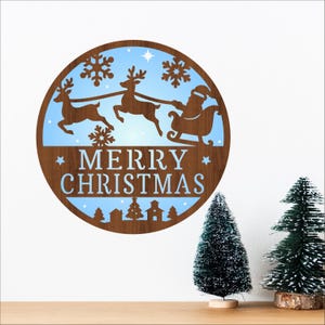 Puede incluir: Decoración navideña redonda de madera con fondo azul que representa a Papá Noel en un trineo tirado por renos, copos de nieve y la frase "MERRY CHRISTMAS". Hay árboles decorativos en primer plano.